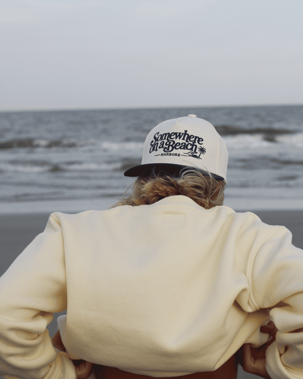 A person seen from behind wearing a cream sweatshirt and a white baseball cap with black 'Somewhere on a Beach' text, standing on a beach with the ocean in the background.