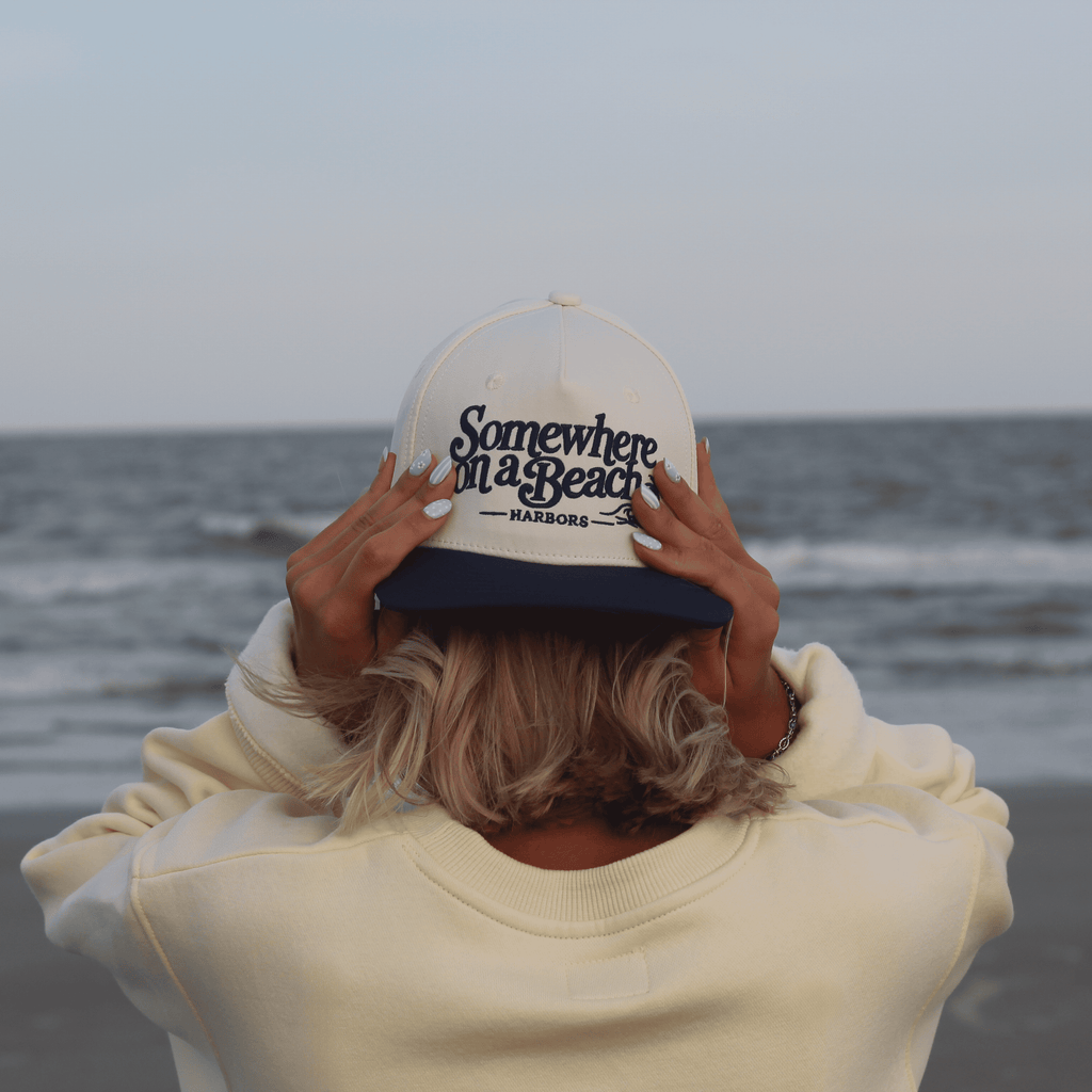 A person standing on a beach with their back to the camera, wearing a cream sweatshirt and a white and navy blue trucker hat with the text 'Somewhere on a Beach - HARBORS' embroidered on the front.