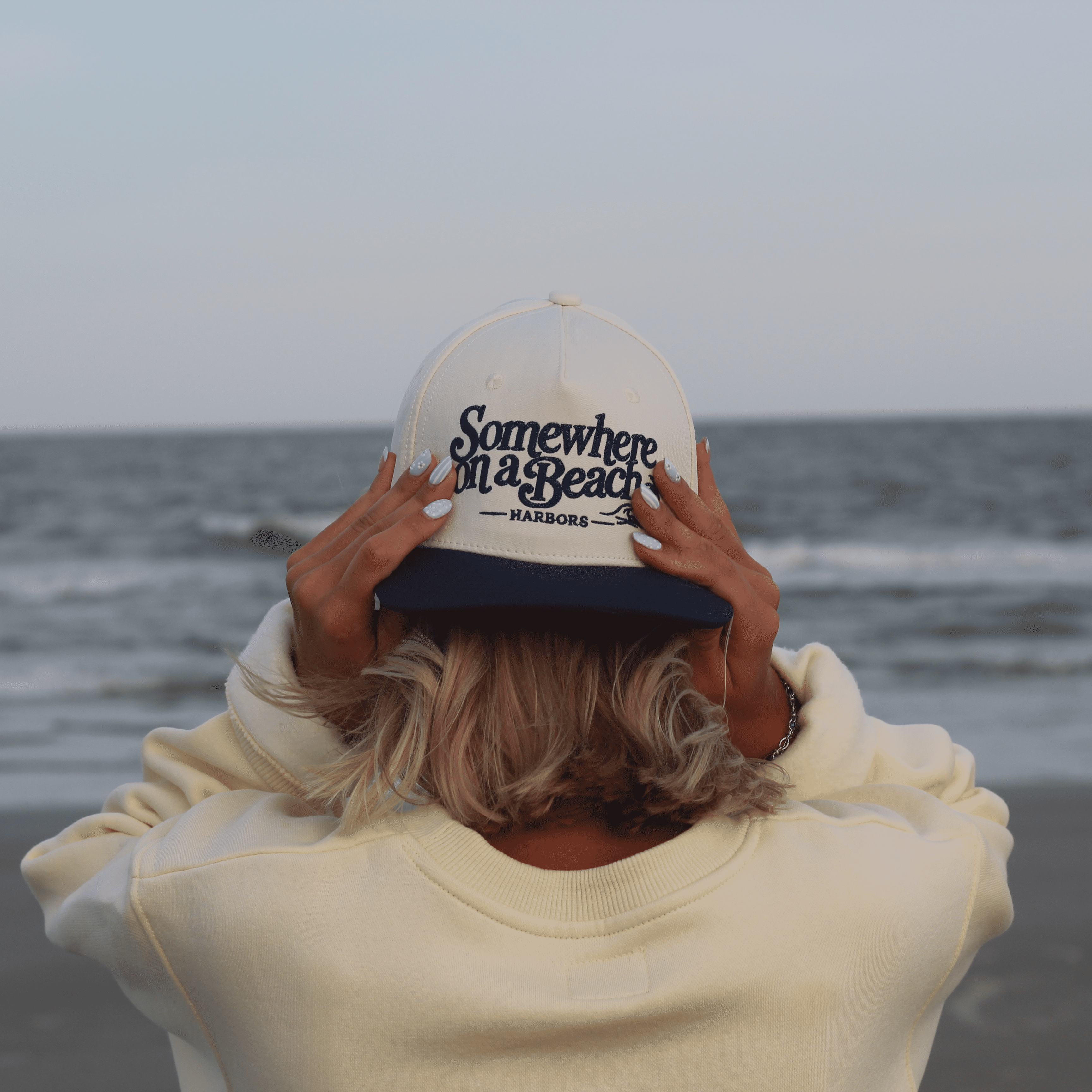 A person standing on a beach with their back to the camera, wearing a cream sweatshirt and a white and navy blue trucker hat with the text 'Somewhere on a Beach - HARBORS' embroidered on the front.