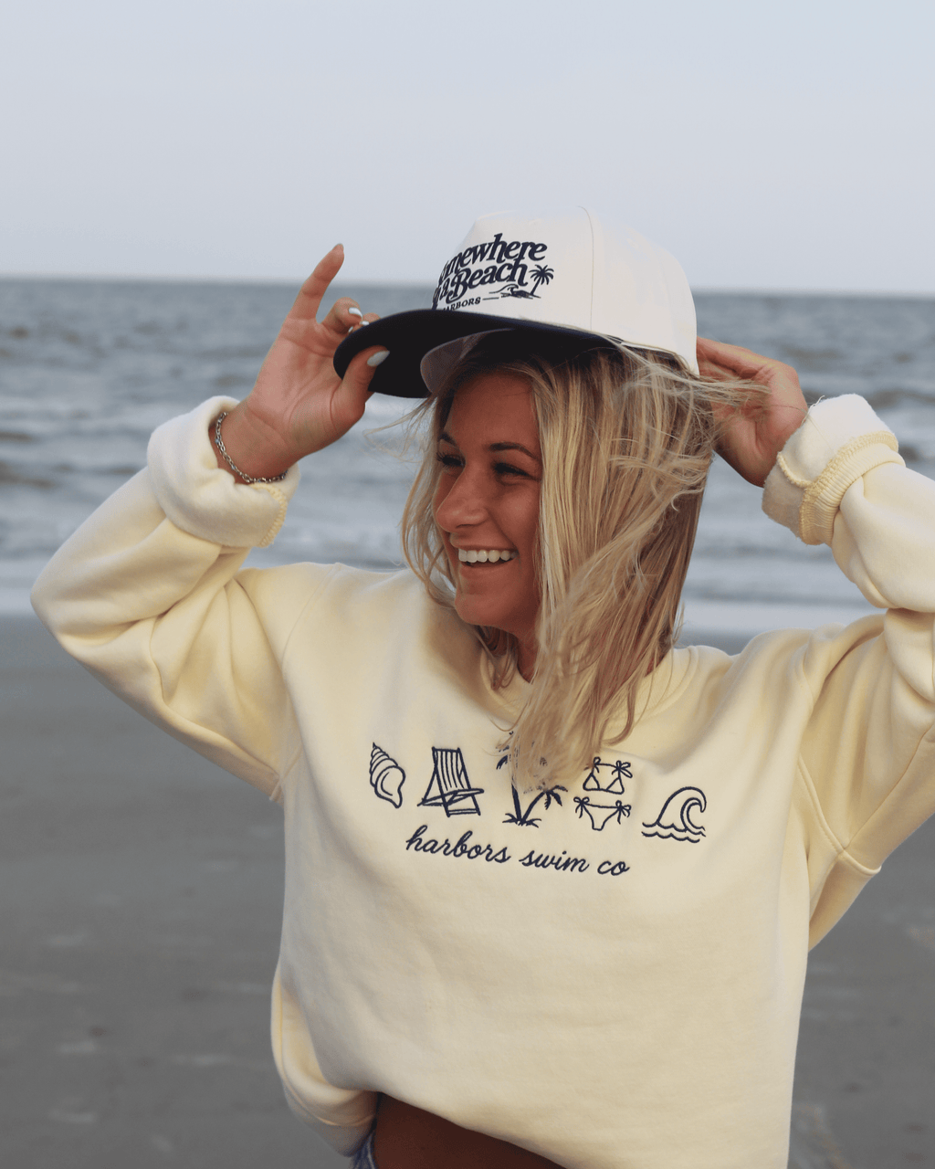 A woman on a beach wearing a cream-colored hoodie with navy line art graphics and the text 'harbors swim co' on the front, along with a matching cream and navy trucker hat.