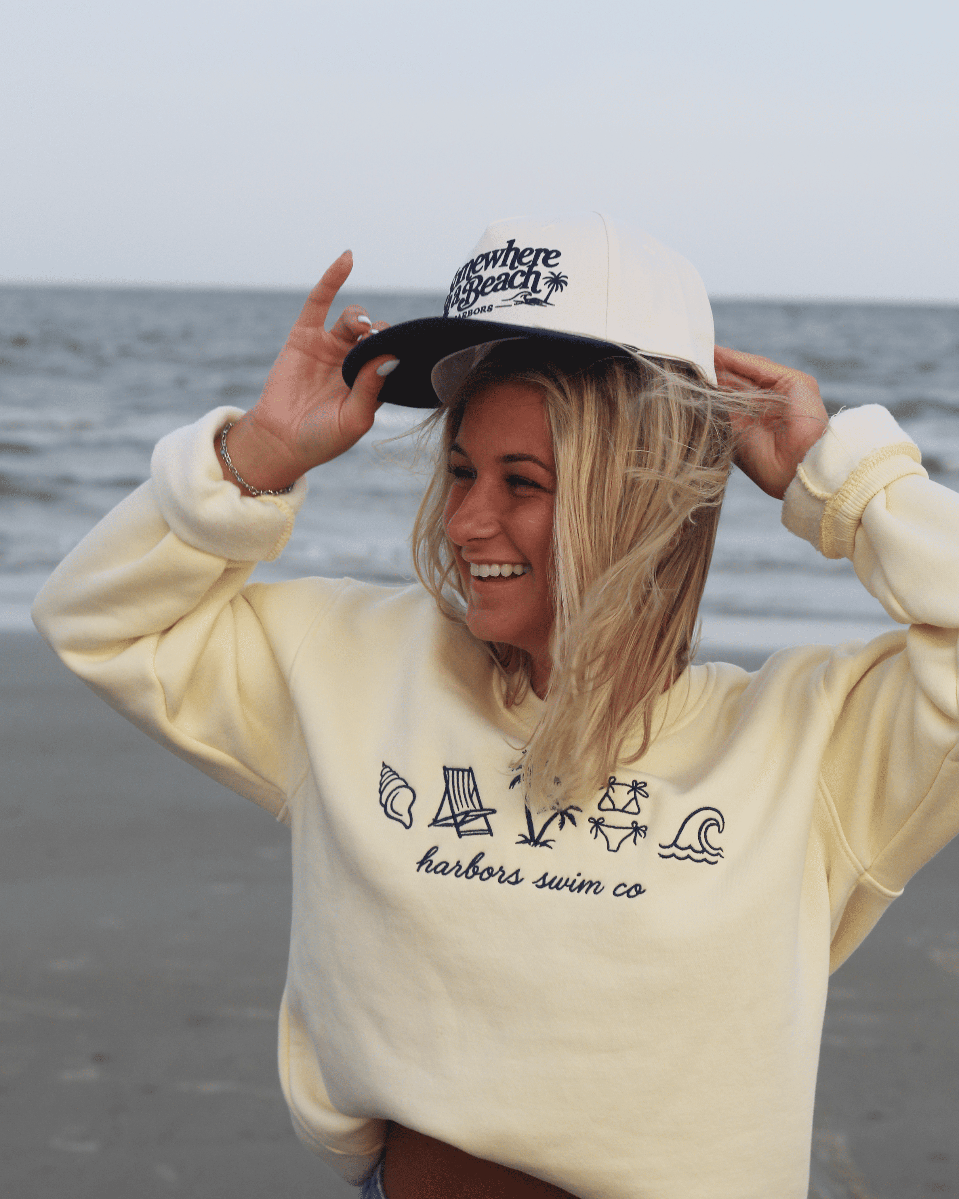 A woman on a beach wearing a cream-colored hoodie with navy line art graphics and the text 'harbors swim co' on the front, along with a matching cream and navy trucker hat.